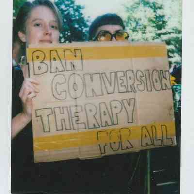 Two people hold a sign that reads "Ban Conversion Therapy For All" at an outdoor event.
