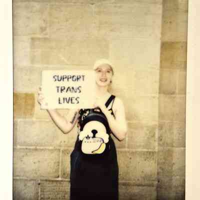 A polaroid of a person smiling holds a supportive sign reading "SUPPORT TRANS LIVES" against a stone wall background.