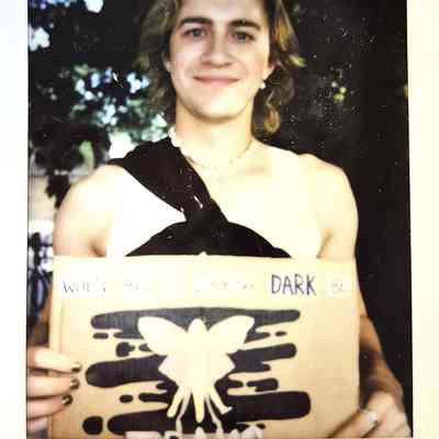 A smiling person holds a sign with an inspirational message, featuring a butterfly design, surrounded by trees.