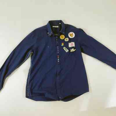 A dark blue button up shirt, with some colourful buttons. It has distinctive 'X' stitching up the side seams and down the arm seams. There are several patches sewn onto the left breast.