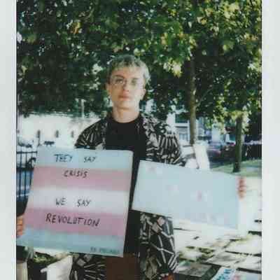 A person stands holding signs, one saying "THEY SAY CRISIS, WE SAY REVOLUTION".