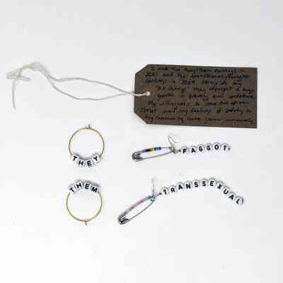 The image shows handmade earrings with pronouns and labels alongside a handwritten note attached with string.