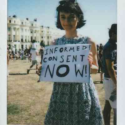 A person holds a sign that reads "Informed Consent Now!" at an outdoor gathering.
