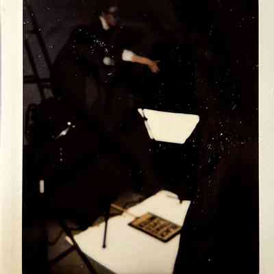 A dark lit polaroid which vaguely shows a photography lighting set up with a placard on the floor and someone stood over it.