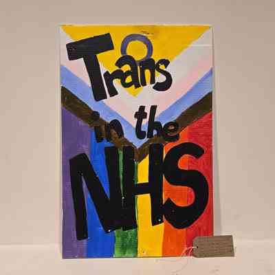 A placard which says “Trans in the NHS” with intertwined flag colors and bold lettering.