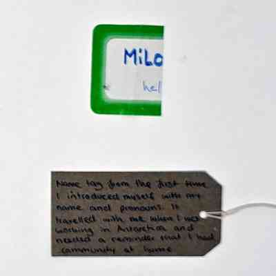 A folded over name label, white with a green border. The name "Milo" is visible, along with "he/". With a brown, handwritten luggage tag.
