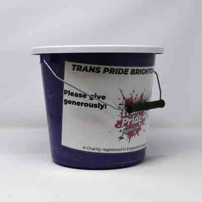 Purple coin collection bucket with white lid and white sticker wrapping around most of body. Several red and purple stickers are pasted on the lid and side with the "Trans Pride Brighton & Hove" logo.
