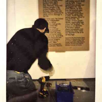 A person wearing a cap fixes something on the floor near a wall with text.