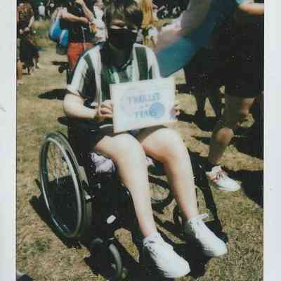 A person in a wheelchair holds a sign saying "THRILLED TRANS," surrounded by people outside.