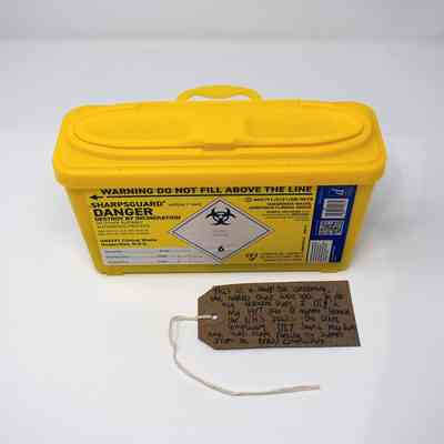 A bright yellow sharps box with a brown, handwritten luggage tag.