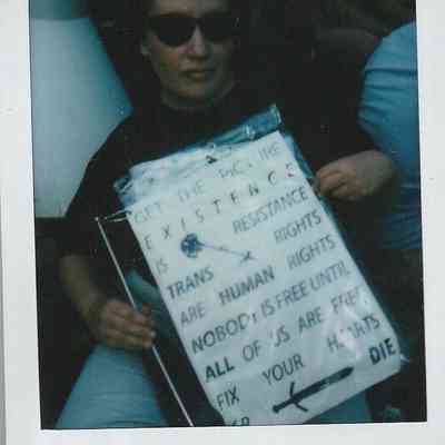 A person wearing sunglasses holds a placard sign which says "Get The Picture Existence Is Resistance Trans Rights Are Human Rights Nobody Is Free Until All Of Us Are Free Fix Your Hearts Or Die".