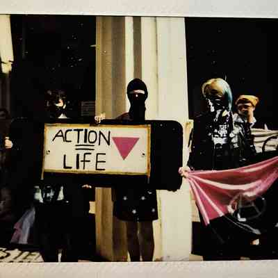 A group of masked individuals hold signs and flags, advocating for "Action = Life".