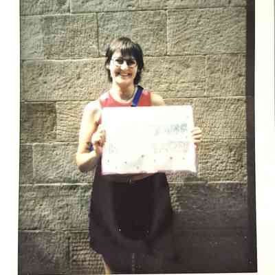 A polaroid of a smiling person holding a colorful sign stands against a stone wall, with text below reading "TP Manchester 24"