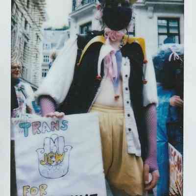 A person in a colorful outfit holds a sign reading "Trans Jew for Palestine" at a public event.
