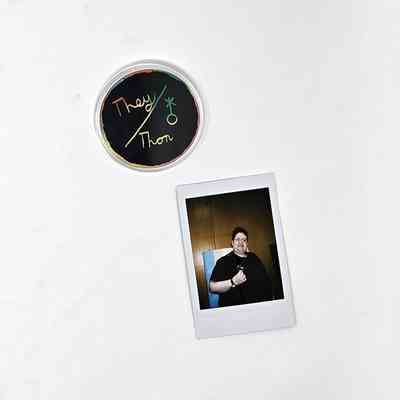 The image displays a round badge with colorful "They/Them" text and a Polaroid
