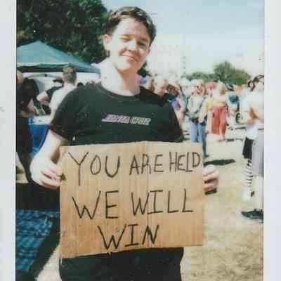 A person is holding a sign reading "YOU ARE HELD WE WILL WIN".