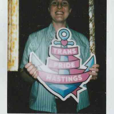 A person smiles and holds a colorful sign reading “Trans Pride Hastings” at Hastings Museum.