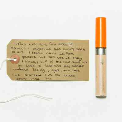 Clear tube of concealer with an orange top. There is a white label on the top left side and faded silver lettering across the bottom half of the tube.