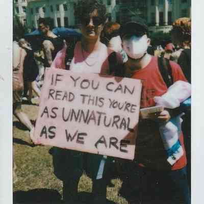 Two individuals at a public event display a sign stating, "If you can read this, you're as unnatural as we are".
