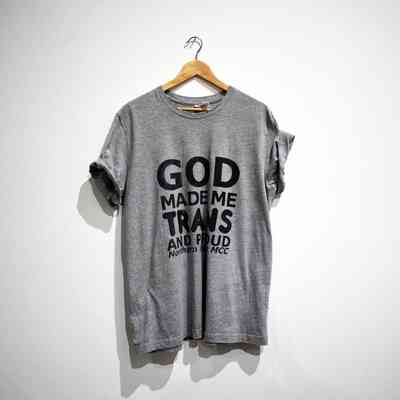 A grey t-shirt on a wooden hanger with text "GOD MADE ME TRANS AND PROUD Northern Lights MCC"