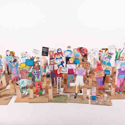 A photograph which is on a white background and has a large group of 'paper people' which are made from cardboard. The people are colourful, wearing different outfits and an array of expression. All of them are holding placards which say different phrases such as 'Trans Kids Deserve Your Love' and 'Queer Love'.