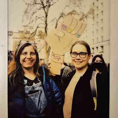 Two smiling individuals hold a paper fist sign with colorful nails in a park during a public event.