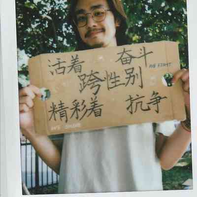 A person holding a cardboard sign with handwritten text, standing outdoors near green leaves.