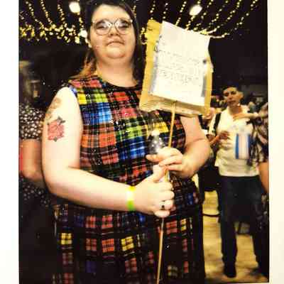 A person in a colorful plaid dress holds a sign in a festive, string-lit venue.