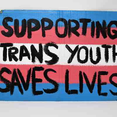 A placard which is painted in trans flag colours of white, pink and blue. Over it has in black paint 'Supporting Trans Youth Saves Lives'.