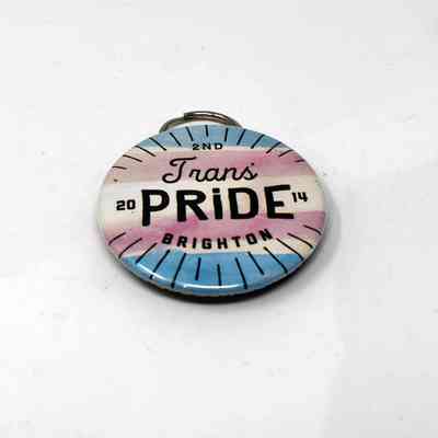 Bottle opener, metal, made to look like a badge reading "2nd Trans Pride Brighton 2014". Blue, white, pink with attached ring on reverse.