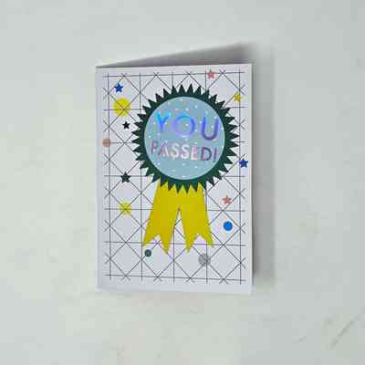 The greetings card that says "YOU PASSED!" inside a blue and green rosette with a yellow ribbon on a background of geometric lines and coloured stars and circles.