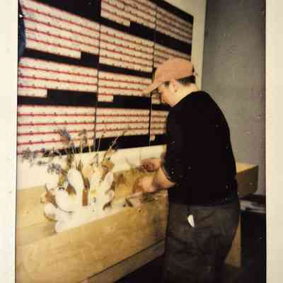 A person wearing a cap is arranging dried flowers on a wooden table in front of a patterned wall.