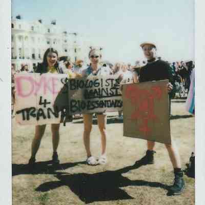 Three people stand with protest signs, gathered for a demonstration outside on a sunny day.