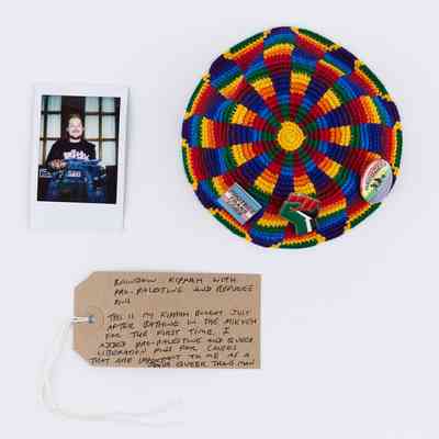 A rainbow coloured Kippah decorated with pro-palestine, pro-refugee, queer and trans pin badges. With a polaroid of the owner and a brown handwritten luggage tag.