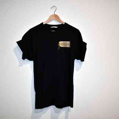 A black T-shirt with rolled sleeves which has the Museum of Transology logo on it hangs on a wooden hanger against a plain wall.
