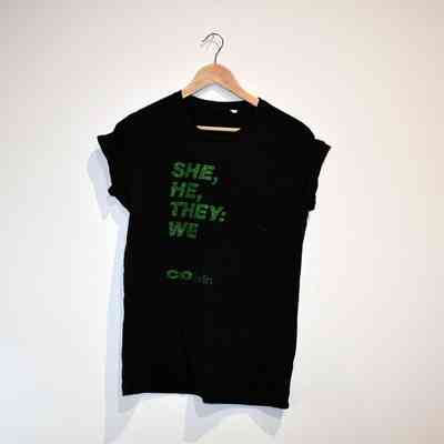 A black T-shirt on a hanger features green text which says 'She, He, They: We'.