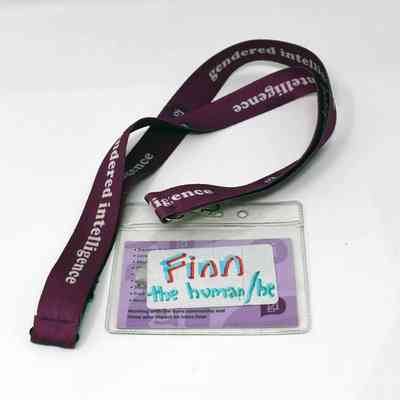 "Gendered intelligence" purple lanyard (470mm length) with plastic card holder, this contains a business card for "Finn Greig". The reverse has a sticker on it that says "Finn the human/he" in felt pen. Finn is lead youth worker at G.I.