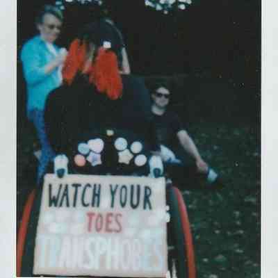 The back of someone sat in a wheelchair and there is a protest sign attached to the wheelchair which says 'Watch your toes transphobes'.