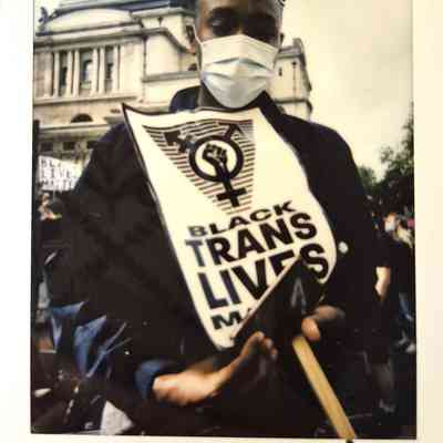 A person wearing a mask holds a "Black Trans Lives Matter" sign in front of a historic building