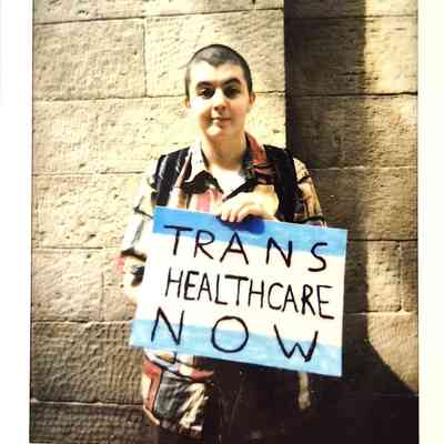 A polaroid of a person holding a sign reading "TRANS HEALTHCARE NOW" stands against a stone wall.