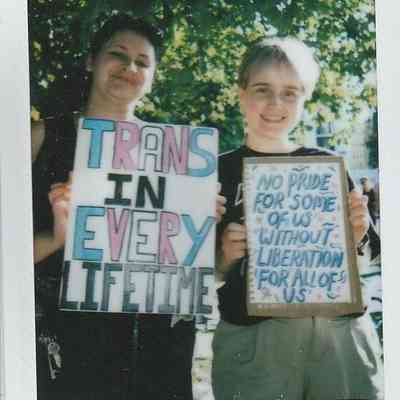 Two people hold signs promoting trans rights and pride, standing outdoors with trees in the background.