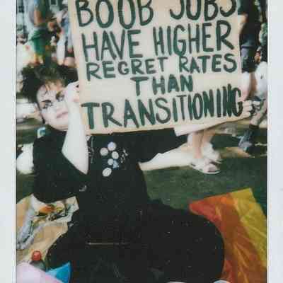 A person is sitting outdoors, holding a sign which says "BOOB JOBS HAVE HIGHER REGRET RATES THAN TRANSITIONING".