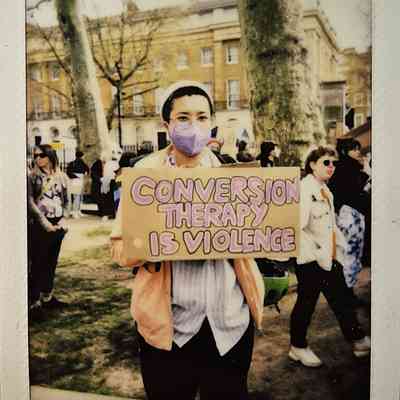 A person in a mask holds a sign saying "Conversion Therapy is Violence" during an outdoor protest.