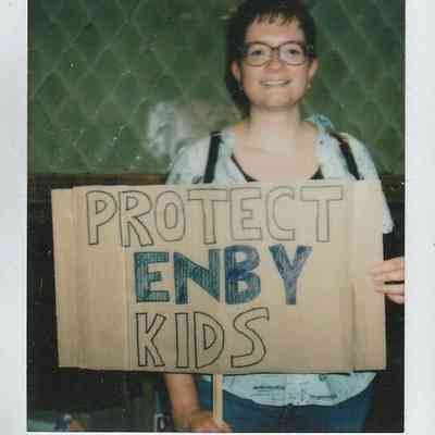 A person is holding a cardboard sign reading "PROTECT ENBY KIDS" with a smile.