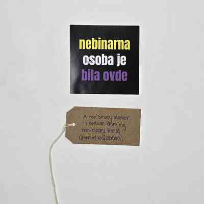 A non-binary sticker in Serbian. It has a black background with text in yellow, white and purple. With a brown, handwritten luggage tag.