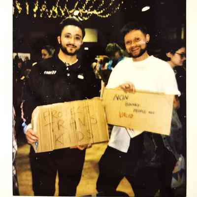 Two people hold signs advocating for trans children and non-binary individuals at an indoor event.
