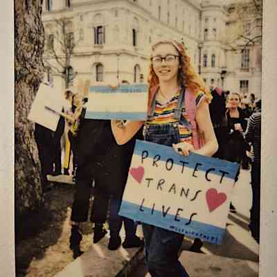 A person wearing colorful clothing holds a "Protect Trans Lives" sign at a rally.
