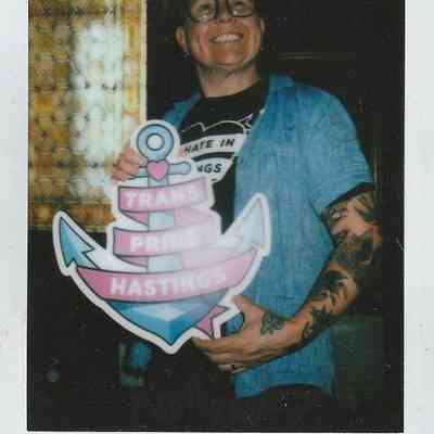 A person smiles while holding a sign that reads "Trans Pride Hastings,".