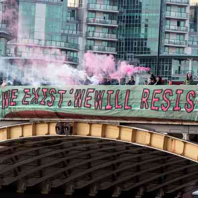 A banner which says 'We Exist: We Will Resist' and is a turquoise colour with the text in pink. It is being hung off a bridge in London with lots of people holding it with others letting off pink smoke. You can see London in the background built up with buildings.