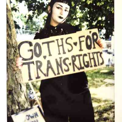 A person stands by a tree holding a sign that says "Goths for Trans Rights" in bold text.